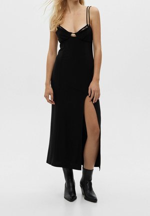 Kobieta nosząca czarną sukienkę bez rękawów z przednim rozcięciem i podwójnymi cienkimi ramiączkami, w połączeniu z czarnymi botkami.