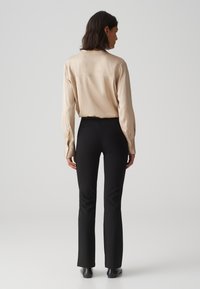 Blouse en soie beige à manches longues avec des poignets boutonnés, assortie à un pantalon noir ajusté avec une légère évasement à l'ourlet, porté avec des chaussures noires.