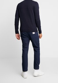 Marineblauer Pullover über gestreiften marineblauen Hosen mit subtiler Textur. Das Outfit wird mit weißen Sneakers vervollständigt. Etikett an den Hosen sichtbar.