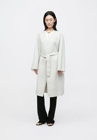 Manteau long en laine gris clair avec manches larges, taille ceinturée et devant ouvert. Associé à un pantalon large noir et des sandales minimalistes.