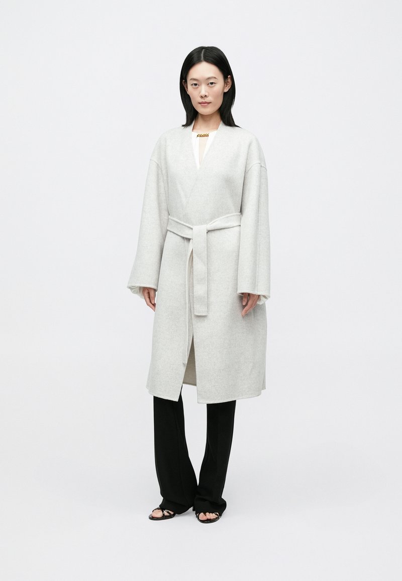 Manteau long en laine gris clair avec manches larges, taille ceinturée et devant ouvert. Associé à un pantalon large noir et des sandales minimalistes.