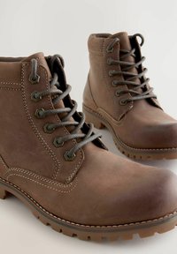 Bruna läderankelboots med grå snören, metallöglor och slitstarka sulor. Har en slät struktur med kontrasterande vit sömnad.