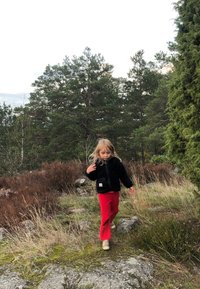 Giacca in pile nera, pantaloni rossi e scarpe beige; camminando su terreno roccioso tra alberi verdi e erba marrone. Capelli biondi lunghi.