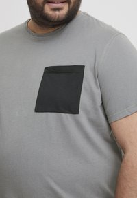 Grijze katoenen T-shirt met een zwarte zak op de linkerborst. Korte mouwen en een ronde halslijn, met contrasterende stiksels.