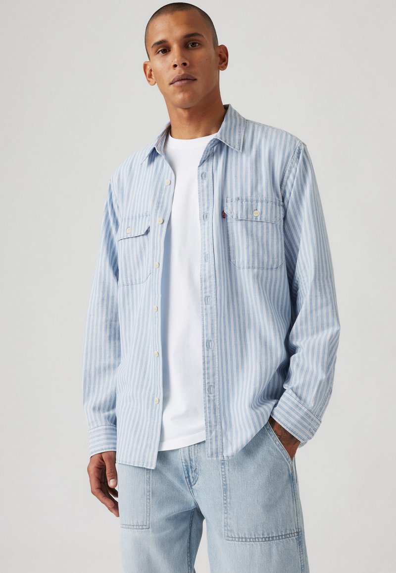Hellblau gestreifte Button-Down-Bluse aus weichem Stoff, mit zwei vorderen Taschen, kombiniert mit einem weißen T-Shirt und hellblauen Jeans.