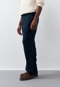 Homme debout portant un pantalon bleu marine, des baskets marron et un pull beige clair, devant un fond uni clair.