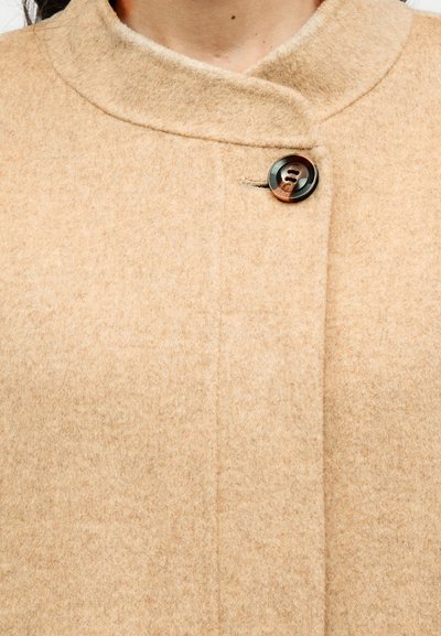 Μπεζ μάλλινο παλτό με ψηλό γιακά, διαθέτοντας μία στρογγυλή κλείσιμο με κουμπί και λεία, απαλή υφή. Μινιμαλιστικός σχεδιασμός και καθαρές γραμμές.