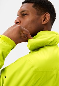 Limoengroene jas met een hoge kraag en een zachte textuur. De kraag heeft een klittenbanddetail en de stof vertoont een subtiel patroon.