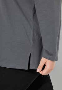 Grijze langemouwen shirt van zachte stof, met zijsplitten aan de zoom. Close-up van de mouw en hand die de stof vasthoudt.