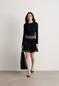 Svart langermet topp parret med et lagdelt svart tyllskjørt, med et stripete elastisk midjebånd. Modellen holder en svart tote bag.