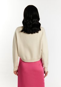 Donna con capelli neri ondulati che indossa un maglione color crema e una gonna di raso rosa brillante, vista di spalle su uno sfondo bianco.