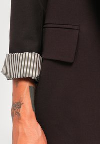 Ylös kääritty tummanruskea takin hiha, jossa on valko-ruskea raidoitus vuorena, paljastaen forearmen tatuoinnin, jossa on abstrakteja mustia kuvioita.