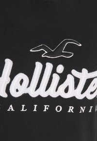 Maglietta nera con testo ricamato bianco "Hollister California" e una grafica di gabbiano sopra. Tessuto morbido con una superficie liscia.