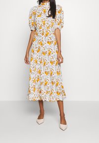 Femme portant une robe midi blanche avec des manches bouffantes et un imprimé floral jaune, associée à des escarpins blancs à bout pointu, se tenant contre un fond uni.