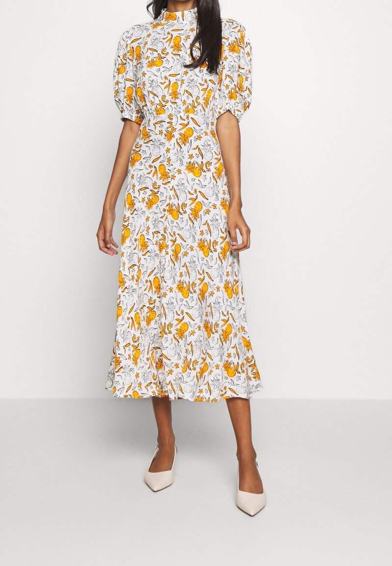 Femme portant une robe midi blanche avec des manches bouffantes et un imprimé floral jaune, associée à des escarpins blancs à bout pointu, se tenant contre un fond uni.