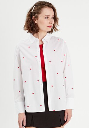 Camicia bianca con pattern di piccoli cuori rossi, maniche lunghe, colletto classico e un tessuto morbido e leggero.