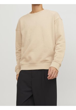 Personne portant un sweat-shirt beige à manches longues, col rond, avec poignets et ourlet côtelés, associé à un pantalon noir ample.