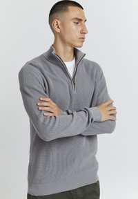 Grauer Zip-Knit-Pullover mit Rippstruktur, ausgestattet mit einem halben Reißverschluss, langen Ärmeln und einem enganliegenden Saum. Über einem weißen Hemd getragen.