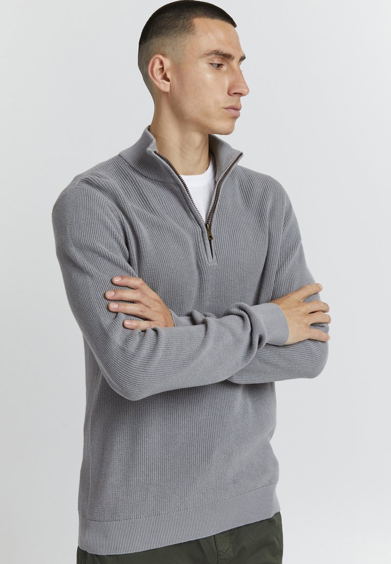 Grauer Zip-Knit-Pullover mit Rippstruktur, ausgestattet mit einem halben Reißverschluss, langen Ärmeln und einem enganliegenden Saum. Über einem weißen Hemd getragen.