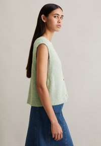 Gilet sans manches en tricot vert clair avec une finition texturée, superposé sur une chemise blanche. Porté avec une jupe en denim. Présente une fermeture par boutons.