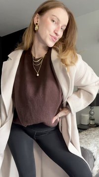 Beige frakke, brun struktureret top og sorte leggings. Guld lagdelte halskæder og store guldøreringe. Blød baggrund med dekorative genstande.