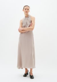 Frau in einem ärmellosen, hellbeigen Halternkleid, mit verschränkten Armen, die schwarze High Heels trägt, vor einem schlichten weißen Hintergrund.
