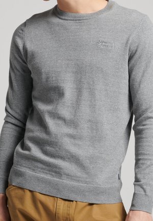 Mann trägt einen hellgrauen Pullover mit Rundhalsausschnitt und dezentem Logo auf der Brust sowie senffarbene Hose, steht vor einfachem Hintergrund.