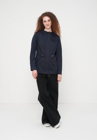 Jeune femme aux cheveux bouclés portant une veste bleu marine, un pantalon noir à jambes larges et des baskets, posant devant un fond blanc.