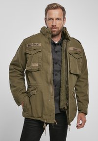 Olive green M65 jacket with a zip front, multiple pockets, and a soft inner lining. Features patches on shoulders; textured fabric.