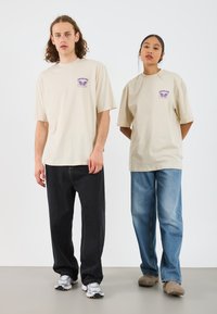 Beige kortermede t-skjorter har et lilla grafisk motiv på venstre bryst. Modellene har på seg mørke jeans og sneakers; én har lyseblå jeans og tresko.