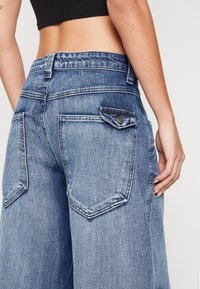 Blå denimjeans med hög midja, vida ben och bakfickor. Har kontraststickning och knappstängning i midjan.