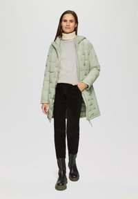 Hellgrüne, gepolsterte Jacke, grauer Pullover, schwarze Hose und schwarze Stiefel mit grünen Sohlen. Komplett mit Reißverschluss, Kapuze und seitlichen Taschen.