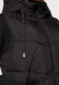 Gilet rembourré noir avec un col haut, présentant un tricot noir texturé sur les côtés et des cordons de serrage ajustables avec un motif réfléchissant.