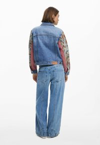 Jeansjack met een blauwe torso, gemusterde mouwen in aardetinten en een leren etiket op de rug. Gecombineerd met wijde denim jeans.