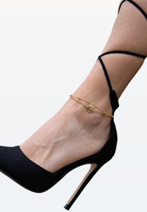 Black suede high heel with a pointed toe and a stiletto heel, featuring a delicate gold chain anklet with a circular charm.