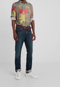 Färggshirt med ett blandat mönster av grafik och text, parat med mörkblå jeans och vita sneakers. Flätat brunt bälte.