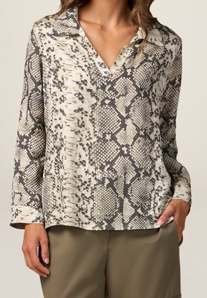 Femme portant un chemisier à manches longues à imprimé serpent avec col, assorti à un pantalon vert olive, debout avec une main dans la poche.