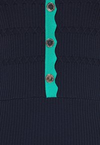 Haut tricoté bleu marine avec une texture côtelée. Il présente un panneau vert festonné et des boutons argentés, ajoutant contraste et détails.