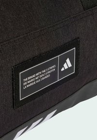 Musta kangaskassi, jossa on teksturoidun pinnan lisäksi suorakulmainen etiketti, jossa on valkoinen teksti ja kolme raitaa logo alareunassa.