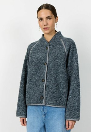 Sieviete, tērpusies tumši pelēkā, teksturētā pogā uz pogas aizdares žaketē ar gaiši pelēku apmali un gaiši zilās džinsās, stāv pret vienkāršu baltu fonu.