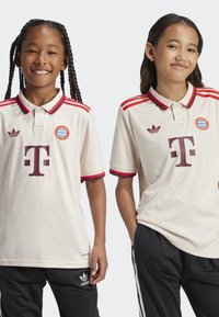 T-shirt crème avec col et manches rayés rouges, arborant des logos d'équipe et un grand motif en maron, associé à un pantalon de sport noir.