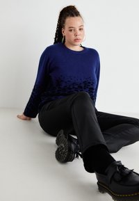 Blauwe trui met een gradientpatroon, zwarte broek en zwarte schoenen. Zachte textuur met ribbelmanchetten en een relaxte pasvorm. Eenvoudig ontwerp van het schoeisel.