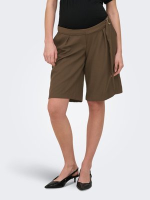 Femeie care poartă pantaloni scurți de culoare verde măsliniu, până la genunchi, cu detaliu de curea laterală și pantofi slingback negri cu vârf ascuțit, stând în picioare pe un fundal simplu.