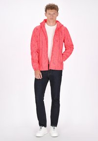 Červená bundá s vzorom a kapucňou, so zipsom a elastickými manžetami, v kombinácii s bielou košeľou, tmavými džínami a bielymi teniskami.