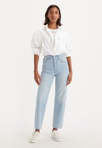 Mulher em pé, vestindo uma camisa branca larga, jeans de cintura alta azul claro, tênis brancos, colar de ouro e pulseira, contra um fundo simples.