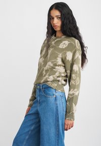 Alyvų žalias megztinis su gėlių raštu, graužtų rankogalių ir apačios. Derinamas su aukšto liemens mėlynomis džinsomis, laisvo kirpimo.