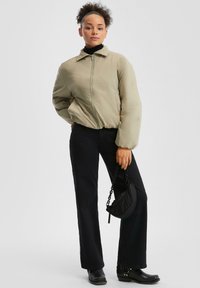Femme aux cheveux bouclés portant une veste zippée beige, un pantalon noir, des bottes noires, et tenant un petit sac à main noir avec une poignée en chaîne.