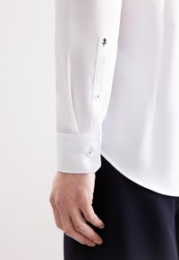 Camicia bianca a texture con polsini aderenti, dotata di chiusura con bottoni e un piccolo dettaglio ricamato a forma di fiore sulla manica. Una mano è visibile accanto alla camicia.