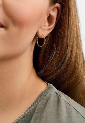 Close-up of a woman’s ear wearing a small gold hoop earring, with light brown hair and a gray top visible.