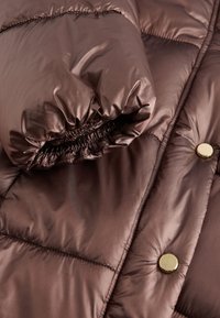 Braune wattierte Jacke mit glänzender, gesteppter Textur und goldenen Beschlägen. Mit elastischem Bündchen und rundem Kragen. Nahaufnahme.